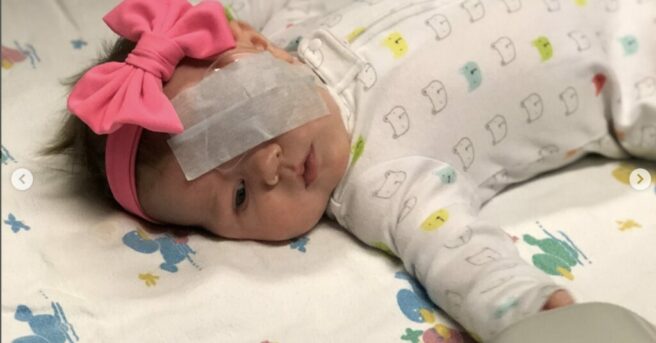 A baby laying with a pink bow and eye patch.