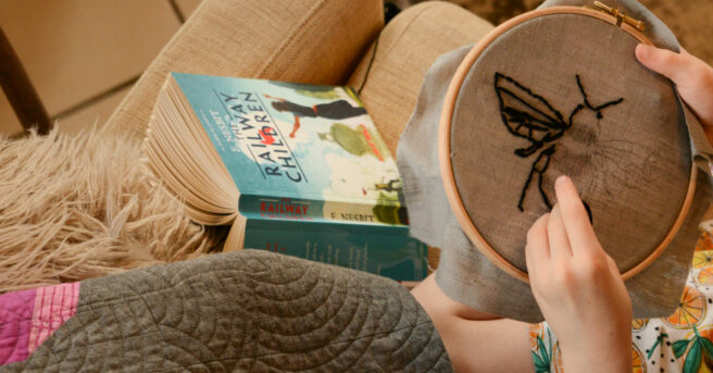 image of a child sitting on a sofa with a copy of The Railway Children cracked open with the pages down and the spine and cover facing up. The child has switched from reading to working on an embroidery hoop with in the pattern of a bee.