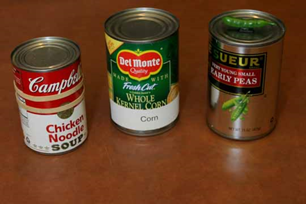 Can of Chicken noodle soup with 2 rubber bands wrapped around it, Corn or corn with white corn label and can or peas with actual pea on top of can. 