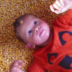 A little boy wearing a red shirt laying on his back looking up at the camera, playing with corn kernels 