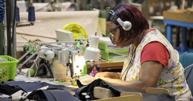 Woman with glasses and headphones sitting at a sewing machine in a factory setting.