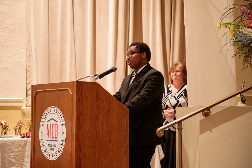 A teenager standing at a podium giving a speech with a teacher behind standing proudly.
