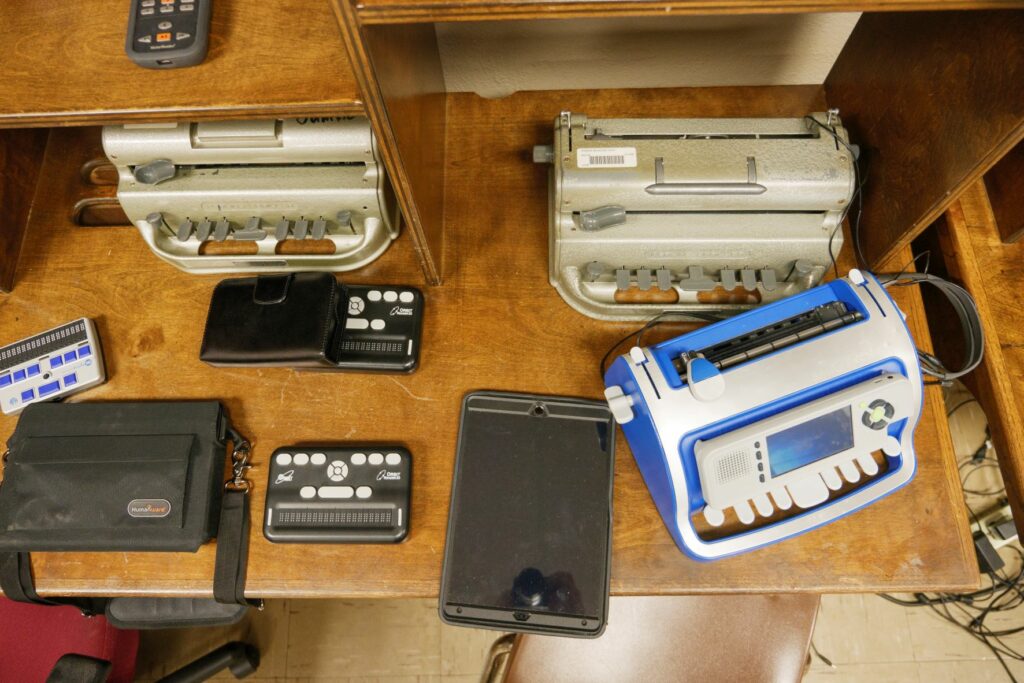 Several Perkins braille writers, a smart brailler and other braille devices displayed on a desk.