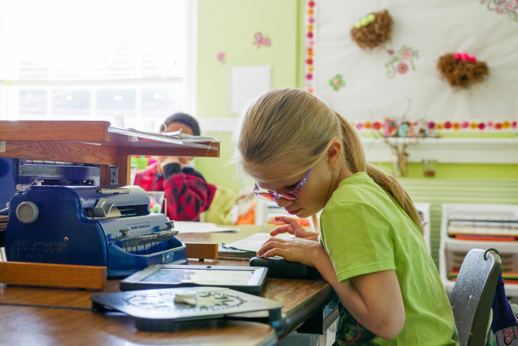 A girl wearing tinted glasses seated at a desk with an iPad, Perkins braille writer, a tactile clock and refreshable braille display.