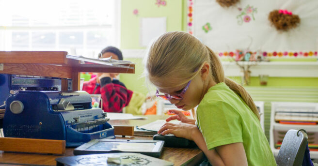A girl wearing tinted glasses seated at a desk with an iPad, Perkins braille writer, a tactile clock and refreshable braille display.