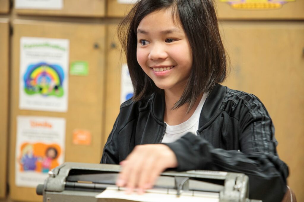 A girl sitting in a classroom using a Perkins Brailler felling what she has written on the paper.