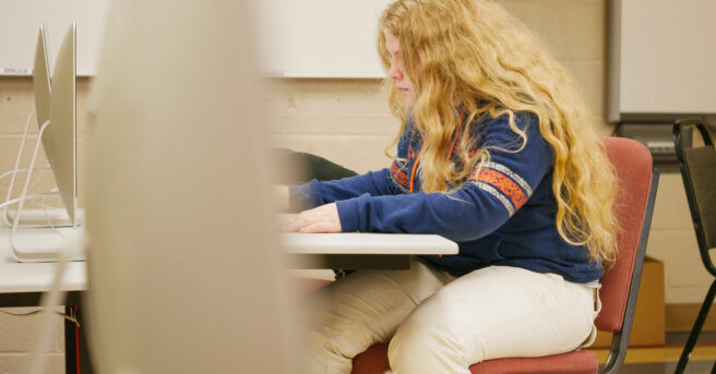 A student sitting at a desk working on a computer in the background.