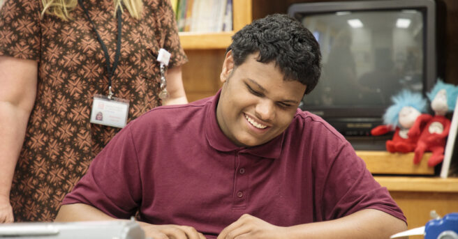 Student in maroon shirt smiling and reading a book.
