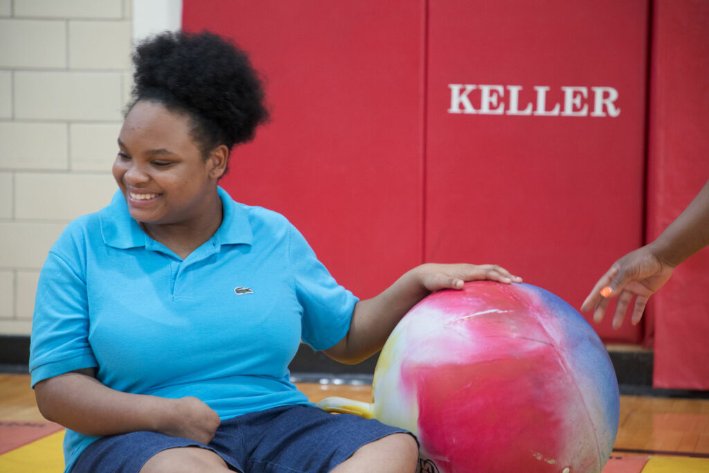 A teen sitting on the gym floor engaging with a large colorful ball.