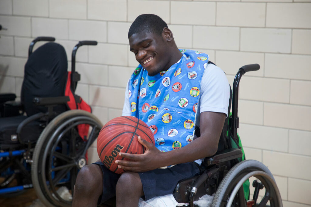 A teen boy in a wheelchair holding and exploring a basketball inside a gym.