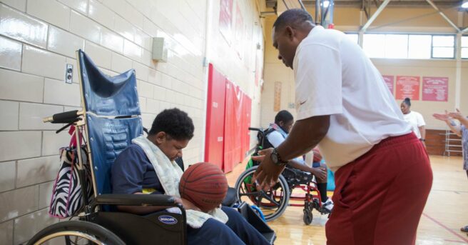 boy in a wheelchair holding a basketball looking down with the teacher holding his hands out.