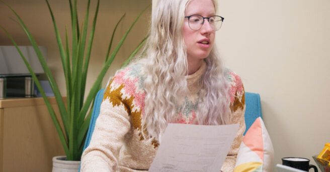 Person with albinism (fair skin, blonde hair, and glasses) sits on a chair and holds a paper.