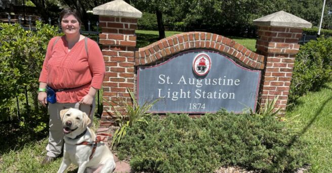 Woman and dog guide in harness stand in front of a sign reading, “St. Augustine Light Station 1874”