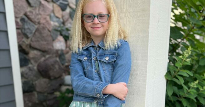 Ally, with light blond hair and teal eyeglasses, clutches her left arm with her right hand