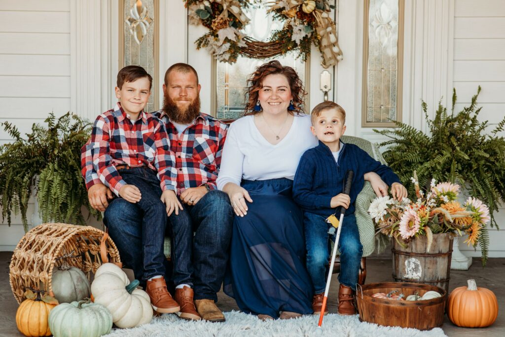 A family photo of Ace, Axel, Ashlyn, and Gunner. Gunner is holding a cane in front of a front door with fall décor.