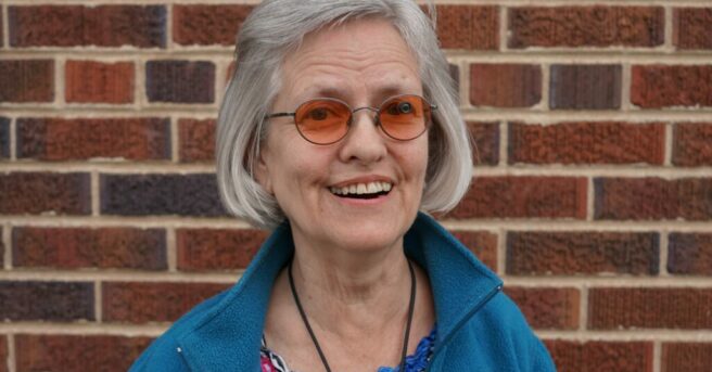 A smiling woman with chin-length gray hair and round orange-tinted glasses. She’s wearing a colorful top and casual jacket.