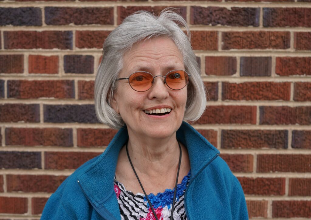 A smiling woman with chin-length gray hair and round orange-tinted glasses. She’s wearing a colorful top and casual jacket.