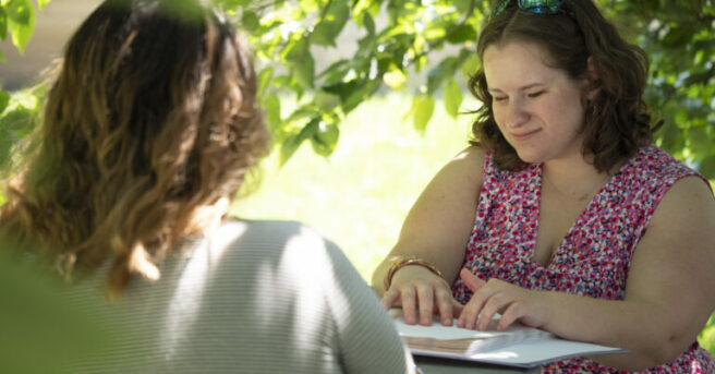 Young woman sitting outside with another woman reading a braille book.