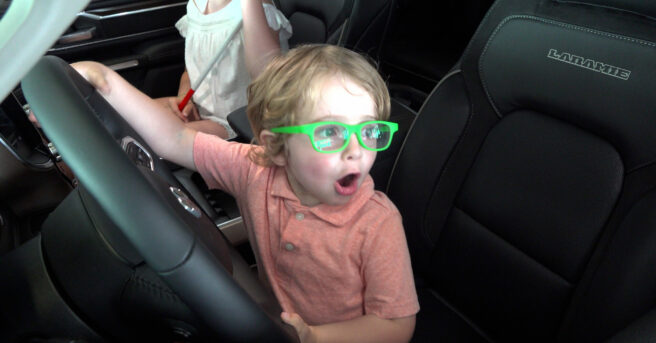 A toddler sitting in a car wearing green glasses holding a steering wheel. 