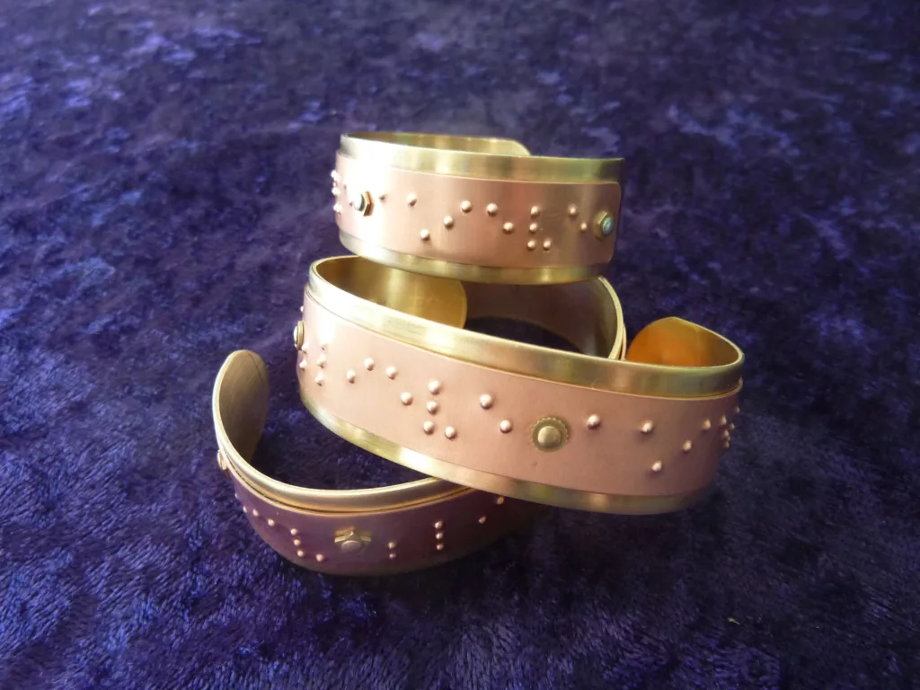 three stacked metal cuff bracelets with braille. top bracelet reads "achieve" available from Elegant Insight jewelry