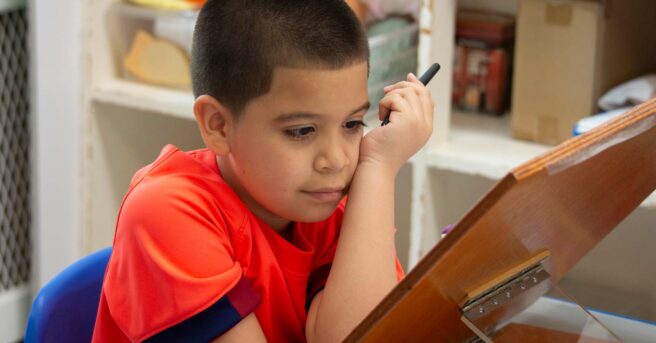 A young boy holding a pen looking at an assignment on a slant board.