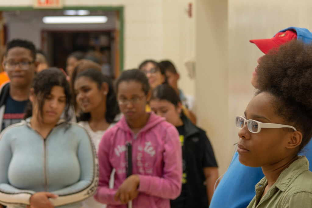 A teenager wearing glasses standing in the hallway with a group of students in the background.
