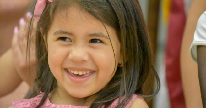 A preschooler smiling wearing a bow in her hair and a dress.