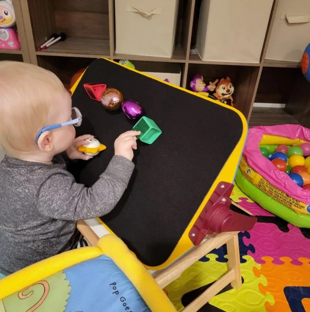Dominic is sitting on the floor and playing with the black felt part of the APH All-In-one board. He is reaching the green square.