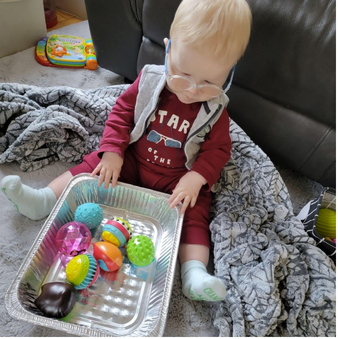 Dominic sitting on the floor with a baking tin filled with various textured balls.