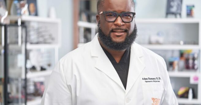 Individual wearing glasses and a white lab coat with embroidered "Adam Ramsey O.D."