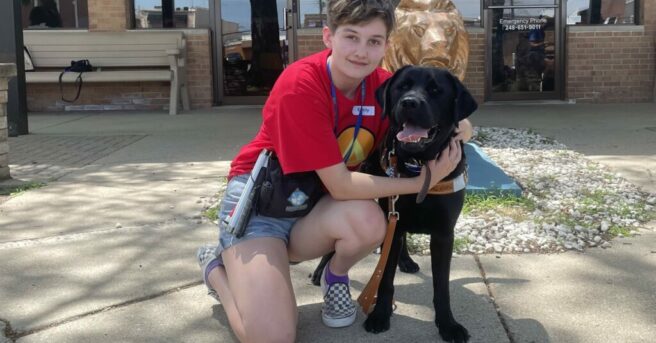 A smiling young woman with short dark hair kneels next to a large black dog, hugging him.