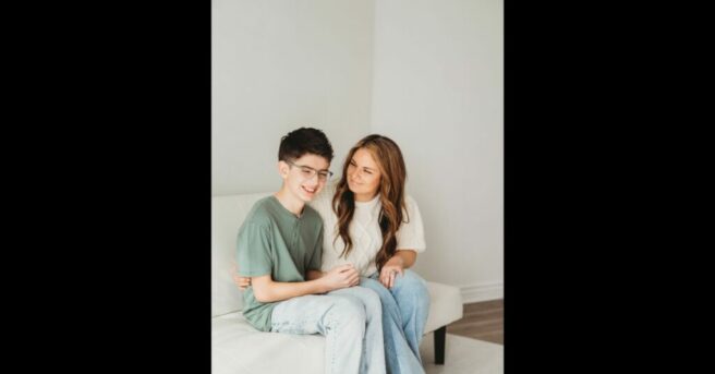 A woman with long dark blonde hair sits with her arm around the waist of a young boy with dark hair and glasses. They are both smiling. (Hilda and her son, Ashton)