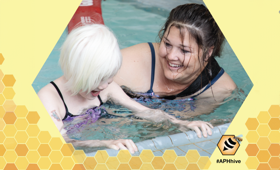 A child in a pool, holding onto the edge with an swim instructor next to the child, with The Hive logo in the lower corner.