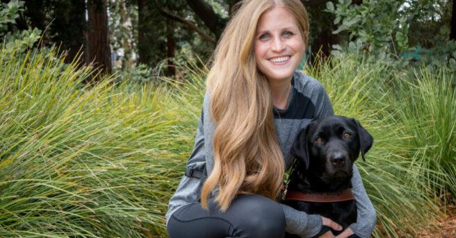 A person in athletic gear smiles outdoors with a dog guide in harness