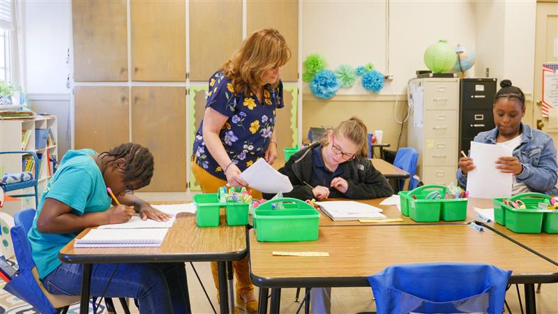 teacher stands beside student wearing eyeglasses in a classroom where students are writing at desks