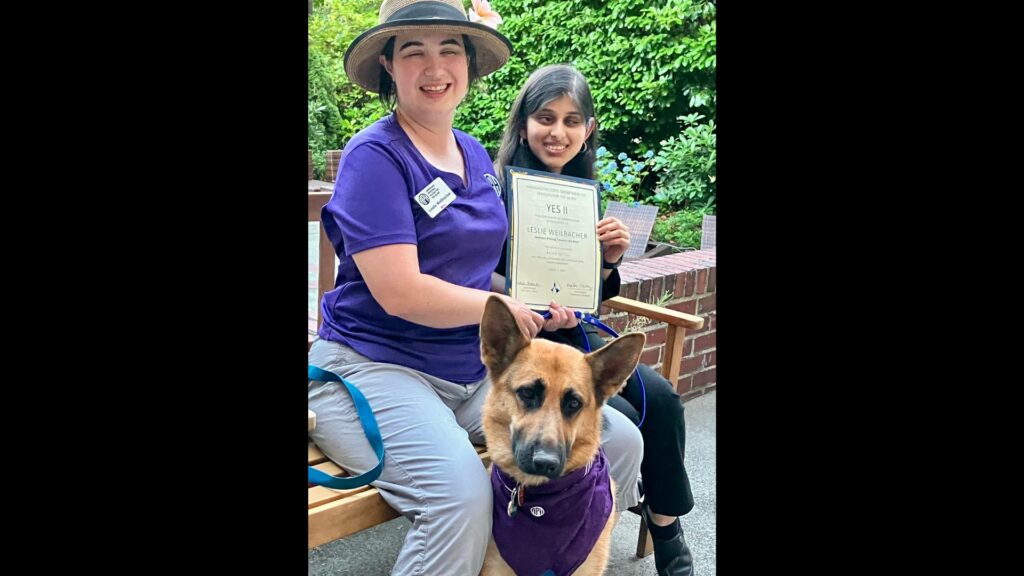 Leslie Weilbacher, Outreach Specialist for the NW Region of APH, is sitting on a bench wearing a purple APH shirt with Arushi next to her. They are holding a certificate of appreciation for the summer. Leslie's German shepherd guide dog, Fame, wears a purple APH bandana.