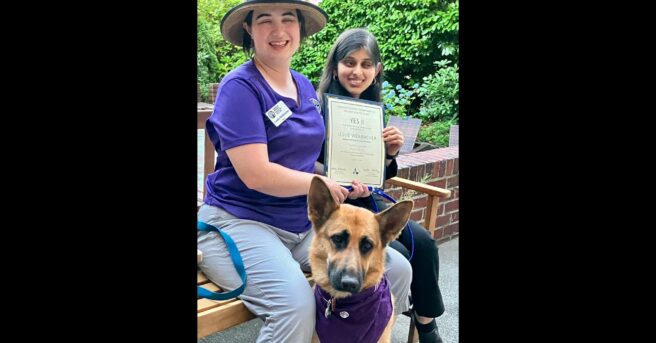 Leslie Weilbacher, Outreach Specialist for the NW Region of APH, is sitting on a bench wearing a purple APH shirt with Arushi next to her. They are holding a certificate of appreciation for the summer. Leslie's German shepherd guide dog, Fame, wears a purple APH bandana.