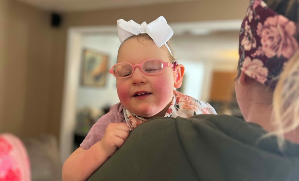 A mom holding a toddler who is wearing pink glasses and a white hair bow smiling.