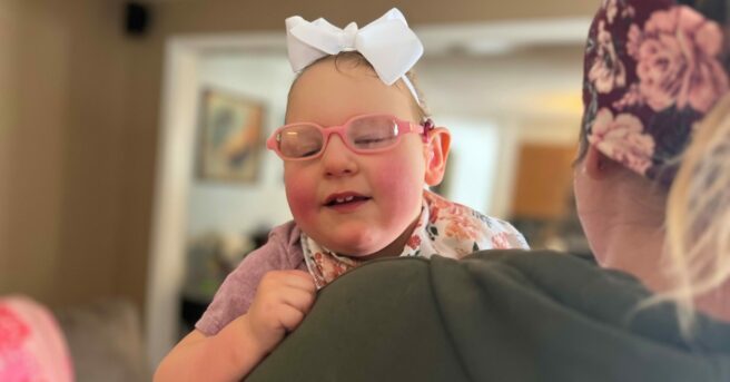 A mom holding a toddler who is wearing pink glasses and a white hair bow smiling.