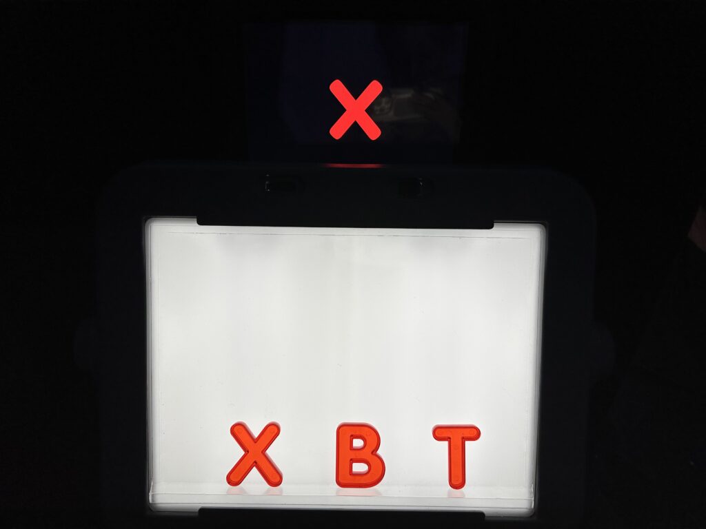 Red, transparent, capital letters X, B, and T sitting on a lightbox with a ledge in a dark room. A red X with a black background displayed on an iPad appears to float behind the lightbox.