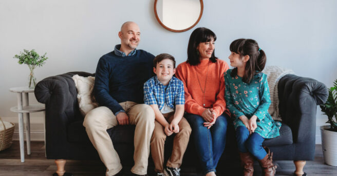 Family of four sit on a couch and look at each other, smiling