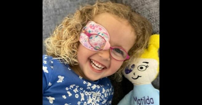 young girl wearing pink eyeglasses and a colorful eyepatch sitting next to a cloth doll with an eye patch and "Matilda" embroidered