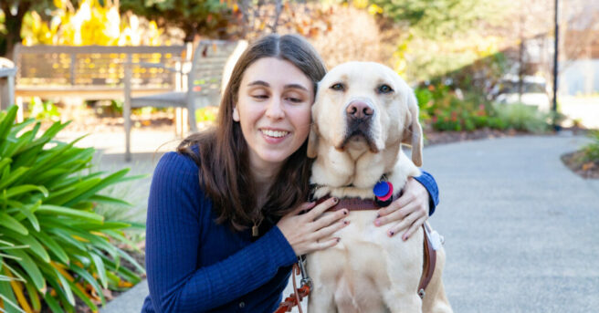 Young adult with her arm around a wheat-colored dog guide in harness