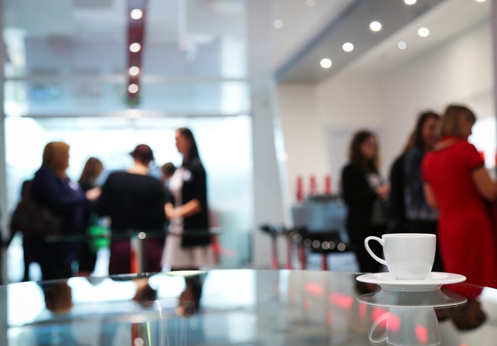 coffee cup resting on a table in the foreground; people in business attire gathering in the background