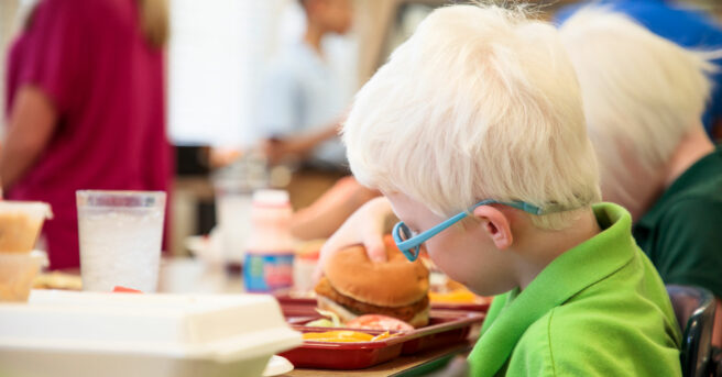 A young boy wearing glasses sitting at a cafeteria table eating.
