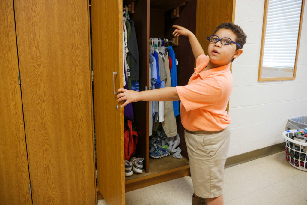 A young boy opening a closet pointing to his clothing. 