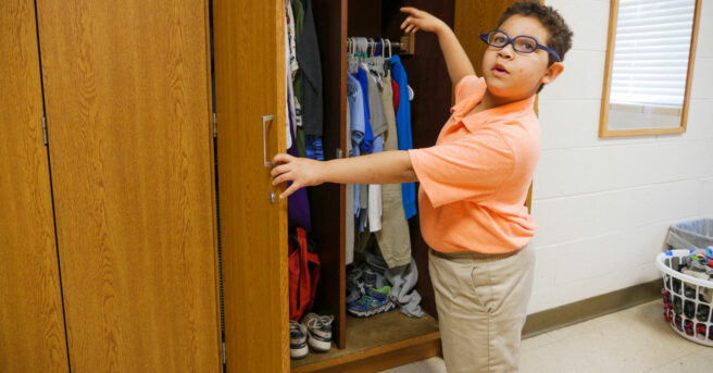 A young boy opening a closet pointing to his clothing. 