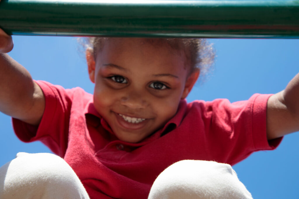 A young child looking through monkey bars smiling outside.