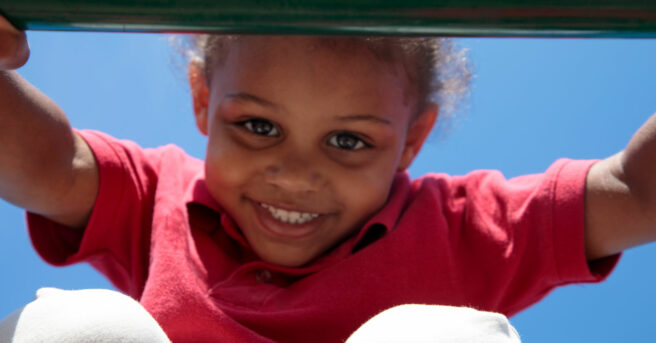 A young child looking through monkey bars smiling outside.