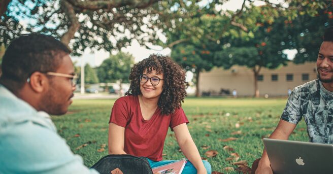 College students sit outside and talk while holding laptops and textbooks.